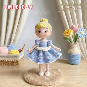 May include: A handmade crochet doll of Cinderella, wearing a light blue dress with white accents. The doll has blonde hair and is standing on a small, round, tan rug. The word "CINDERELLA" is displayed above the doll.