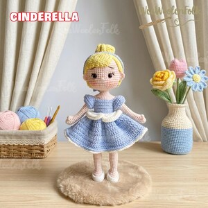 May include: A handmade crochet doll of Cinderella, wearing a light blue dress with white accents. The doll has blonde hair and is standing on a small, round, brown rug. The word "CINDERELLA" is displayed above the doll.