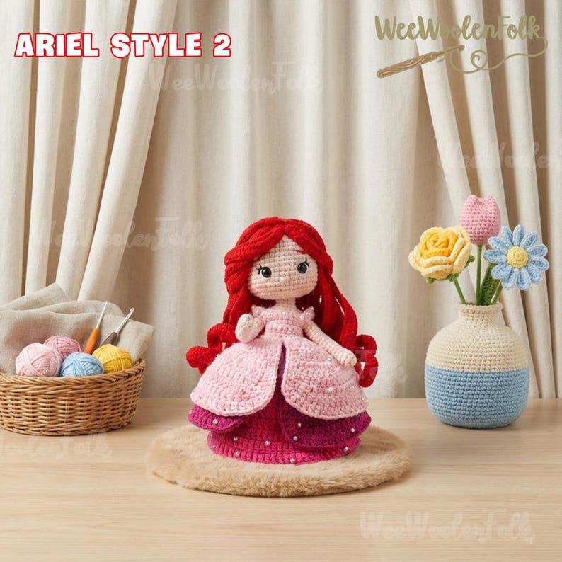 May include: Ariel Style 2 crochet doll with red hair and a pink dress. The doll is handmade and sits on a round, tan surface. A small basket with yarn and a vase with crochet flowers are also in the image.