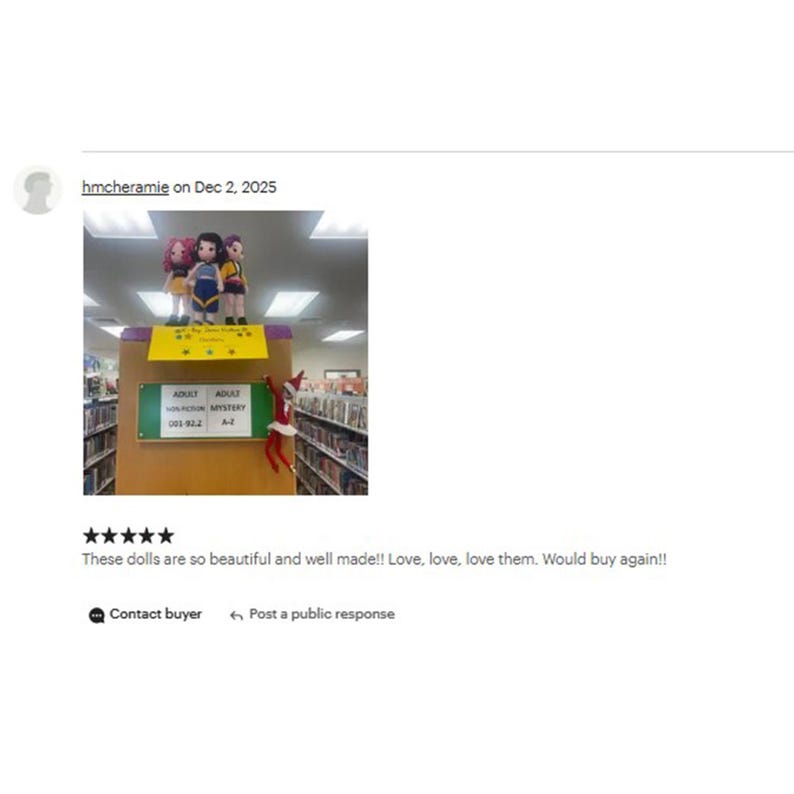 Op de afbeelding: Drie kleine poppen in kleurrijke outfits staan op een boekenplank in een bibliotheek. Op de plank staat "ADULT FICTION MYSTERY". Een elfdecoratie is zichtbaar. De afbeelding bevat een klantbeoordeling.