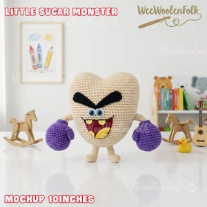 Puede incluir: Un juguete de monstruo en forma de coraz&oacute;n tejido a ganchillo con cuerpo beige, cejas negras y ojos azules. Tiene guantes de boxeo morados y una amplia sonrisa. El texto "LITTLE SUGAR MONSTER" y "MOCKUP 25 cm" son visibles.
