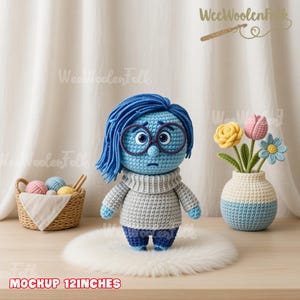 May include: A crocheted doll of a character with blue skin, hair, and glasses, wearing a gray sweater and blue pants. The doll is approximately 12 inches tall. A small basket with yarn balls and a vase with crocheted flowers are also in the image.