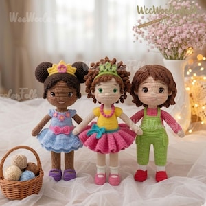 May include: Three handmade crochet dolls with different skin tones and hairstyles. The dolls are wearing colorful outfits, including dresses, tutus, and overalls. A basket of yarn balls sits in the foreground. The dolls are displayed on a white surface.