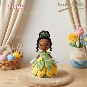 May include: Crocheted doll of Princess Tiana in a green and yellow dress, with a black bun and a green crown. The doll is on a brown surface, with a basket of yarn and a vase of crocheted flowers in the background. The word "TIANA" is in the top left corner.