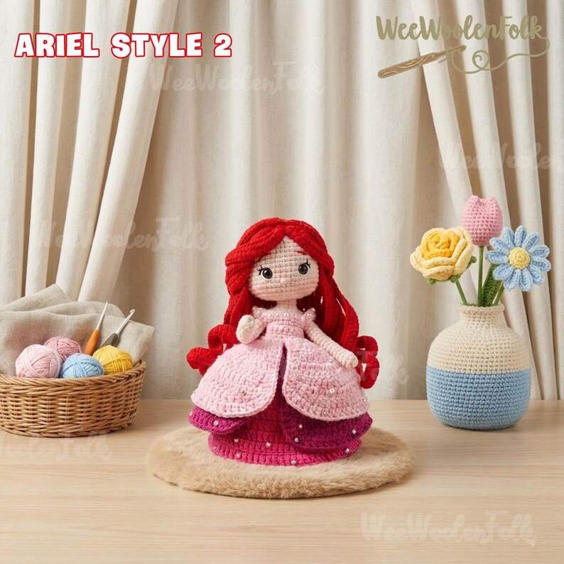 May include: Ariel Style 2 crochet doll with red hair and a pink dress. The doll is displayed on a round, light brown surface. A small basket with yarn and a vase with crochet flowers are also in the image.