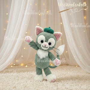 May include: A handmade crochet cat doll in shades of green, white, and pink. The cat wears a green beret and has a white fluffy chest and tail. The doll is standing on a cream-colored rug. The brand name "WeeWoolenFolk" is visible in the background.