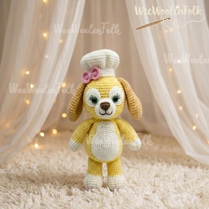 May include: A handmade crochet dog doll wearing a chef's hat and a pink bow. The dog is yellow with white accents on its paws, belly, and hat. The doll has brown eyes and long, floppy ears. The background includes the text "WeeWoolenFolk".