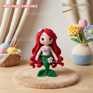 May include: A handmade crochet mermaid doll, approximately 12 inches tall, with vibrant red hair, a lavender top, and a green tail. The doll is displayed on a fluffy, light brown surface. The image also includes a crochet vase with flowers and a basket of yarn.