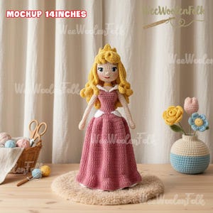 Może przedstawiać: Ręcznie robiona szydełkowa lalka, o wysokości około 35,5 cm, ubrana w r&oacute;żową suknię z białym kołnierzem i złotą koroną. Lalka ma blond włosy i stoi na beżowym dywanie. Widoczny jest tekst "MOCKUP 14 INCHES".