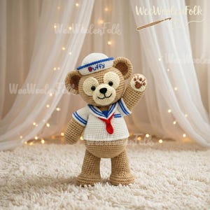 May include: A handmade crochet bear wearing a sailor outfit. The bear is tan with a white and blue sailor shirt, a red tie, and a white hat with "Duffy" on it. The bear is waving with a paw print on its hand.