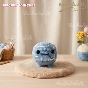 May include: A light blue crocheted sphere-shaped plush toy with a smiling face and small legs. The toy is approximately 10 inches in diameter and sits on a fluffy, light brown rug. A small vase with crocheted flowers is in the background.