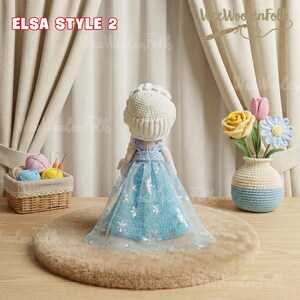 May include: A handmade crochet doll in the style of Elsa from Frozen, wearing a blue dress with silver snowflake accents and a white crown. The doll is displayed on a brown rug, with the text "ELSA STYLE 2" visible. A crochet vase with flowers is in the background.