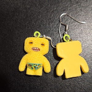 May include: A pair of novelty earrings featuring a yellow figure with a textured surface, detailed with a face and swim trunks. The earrings have silver hooks and green accents. One earring shows the front, the other the back.