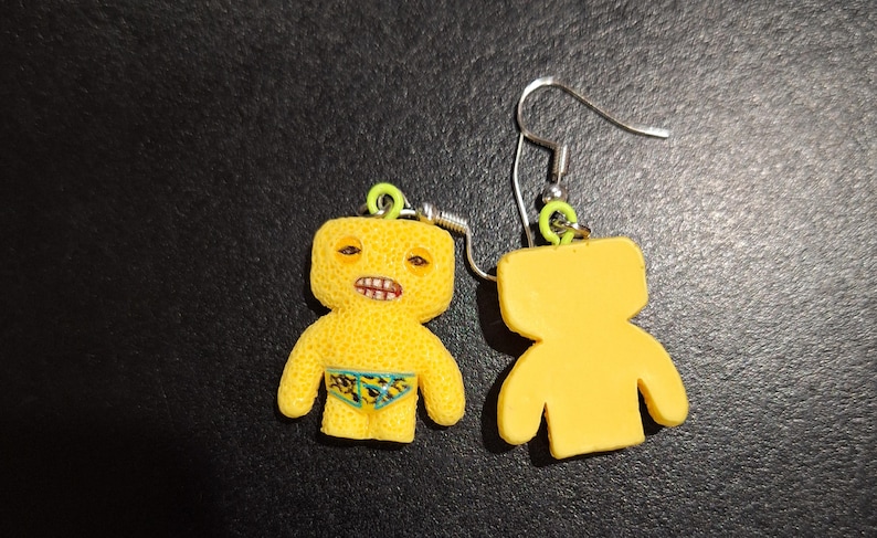 May include: A pair of novelty earrings featuring a yellow figure with a textured surface, detailed with a face and swim trunks. The earrings have silver hooks and green accents. One earring shows the front, the other the back.