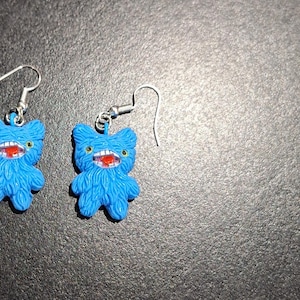 May include: A pair of blue monster-shaped earrings. Each earring features a furry body, a wide red mouth with white teeth, and small yellow eyes. The earrings have silver-colored fish hook closures.