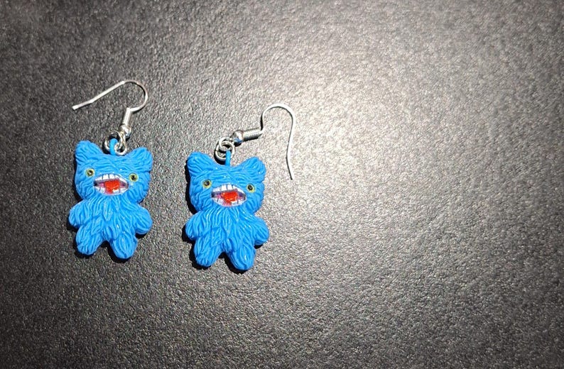 May include: A pair of blue monster-shaped earrings. Each earring features a furry body, a wide red mouth with white teeth, and small yellow eyes. The earrings have silver-colored fish hook closures.