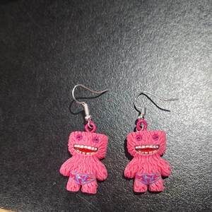 May include: A pair of pink monster-shaped earrings. Each earring features a fuzzy pink body, a wide open mouth with red lips and white teeth, and small blue patterned underwear. The earrings have silver-colored fish hook ear wires.