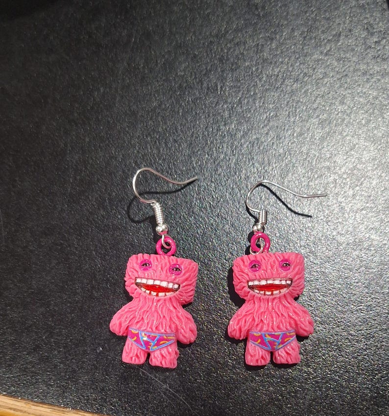 May include: A pair of pink monster-shaped earrings. Each earring features a fuzzy pink body, a wide open mouth with red lips and white teeth, and small blue patterned underwear. The earrings have silver-colored fish hook ear wires.