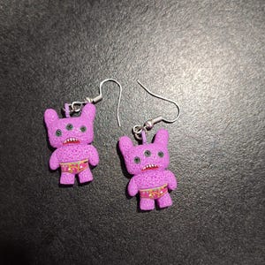 May include: A pair of novelty earrings featuring purple monster figures with three eyes, small arms, and red mouths. The monsters wear patterned underwear. The earrings have silver-colored fish hook closures.