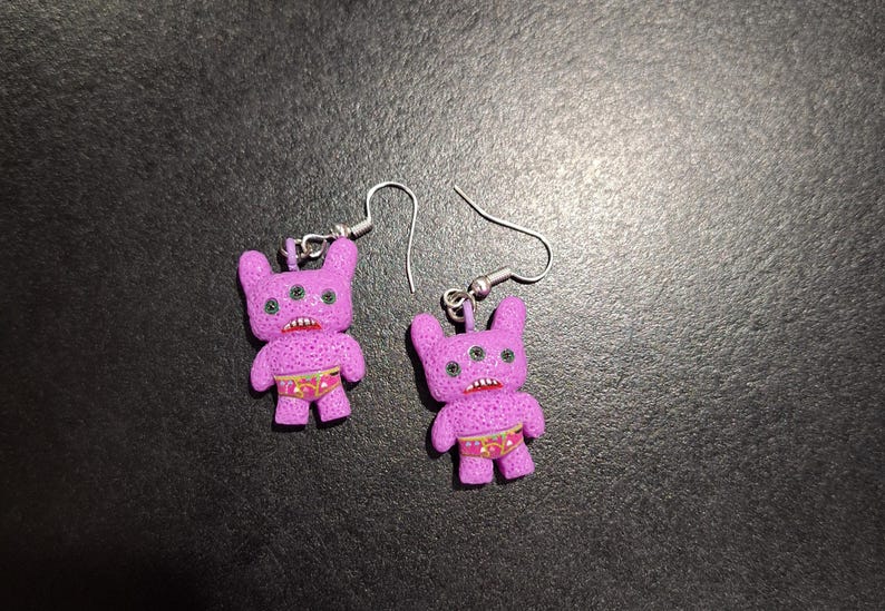 May include: A pair of novelty earrings featuring purple monster figures with three eyes, small arms, and red mouths. The monsters wear patterned underwear. The earrings have silver-colored fish hook closures.
