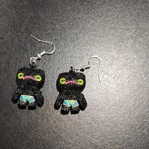 May include: A pair of black novelty earrings featuring a cartoonish figure with yellow eyes, a pink mouth, and blue and yellow patterned shorts. The earrings have silver-colored fish hook closures.