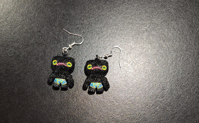 May include: A pair of black novelty earrings featuring a cartoonish figure with yellow eyes, a pink mouth, and blue and yellow patterned shorts. The earrings have silver-colored fish hook closures.