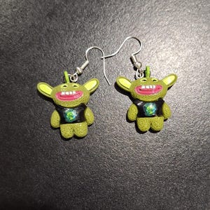 May include: A pair of novelty earrings featuring green cartoon characters with large ears, red mouths, and black shirts. The earrings have silver-colored hooks.