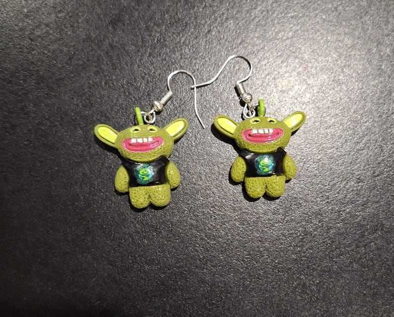 May include: A pair of novelty earrings featuring green cartoon characters with large ears, red mouths, and black shirts. The earrings have silver-colored hooks.