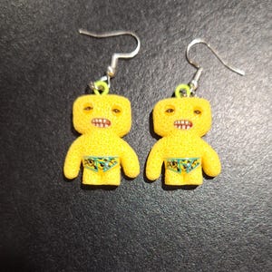 May include: A pair of yellow, cartoon-like earrings. Each earring features a small, humanoid figure with a textured surface, open mouth, and small swim trunks. The earrings have silver-colored hooks.