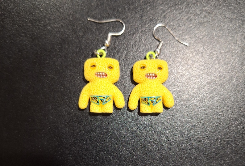 May include: A pair of yellow, cartoon-like earrings. Each earring features a small, humanoid figure with a textured surface, open mouth, and small swim trunks. The earrings have silver-colored hooks.