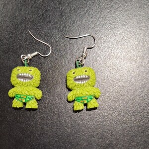 May include: A pair of novelty earrings featuring lime green monster figures with wide, toothy grins and green swim trunks. The earrings have silver-colored fish hook closures and are set against a dark background.