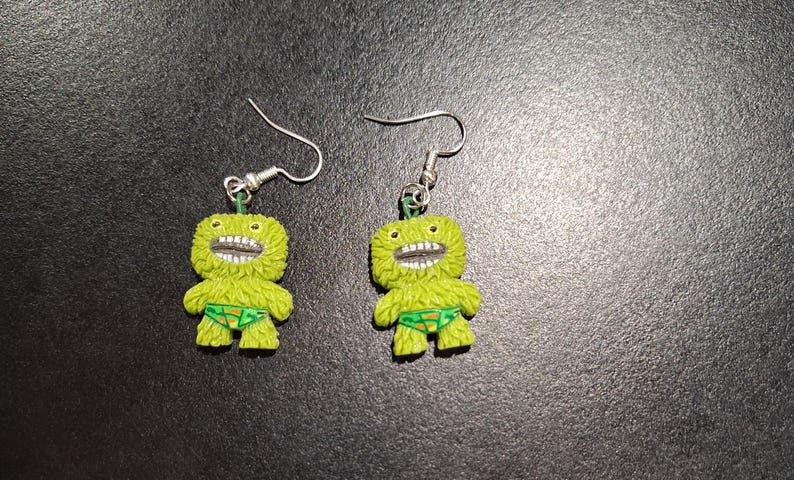 May include: A pair of novelty earrings featuring lime green monster figures with wide, toothy grins and green swim trunks. The earrings have silver-colored fish hook closures and are set against a dark background.
