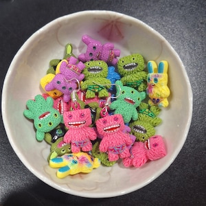May include: A white bowl filled with colorful monster-shaped earrings. The earrings are in various colors, including pink, green, purple, and yellow. Each monster has a unique design, with open mouths and detailed features. The earrings are made of a textured material.