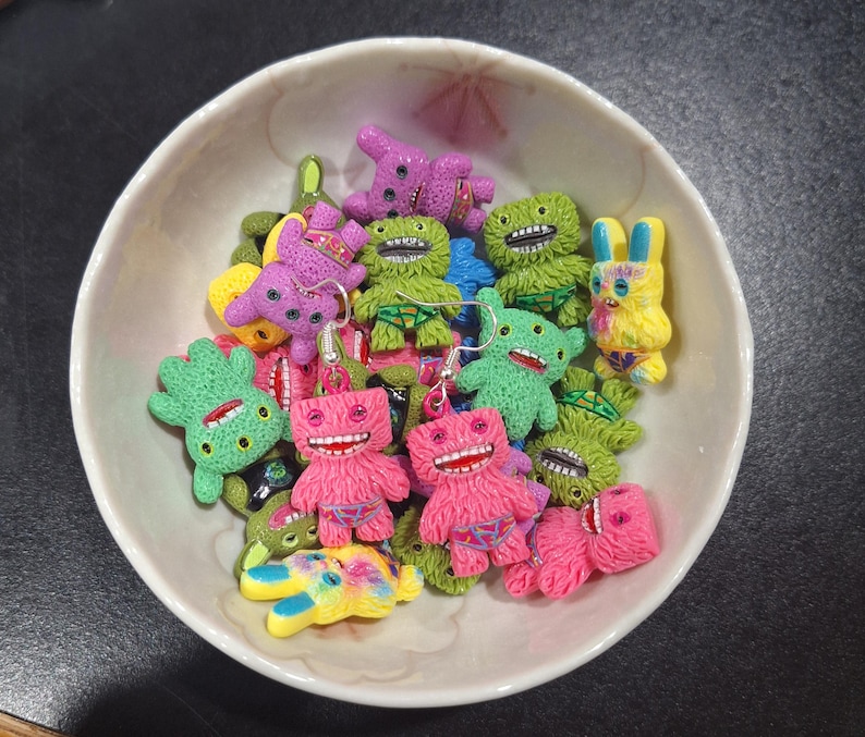 May include: A white bowl filled with colorful monster-shaped earrings. The earrings are in various colors, including pink, green, purple, and yellow. Each monster has a unique design, with open mouths and detailed features. The earrings are made of a textured material.