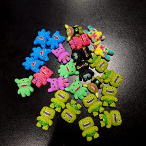 May include: A collection of colorful, miniature monster figurines in various colors, including blue, pink, green, yellow, and black. Each figurine has a unique design with open mouths and small details. The figurines are scattered on a dark surface.