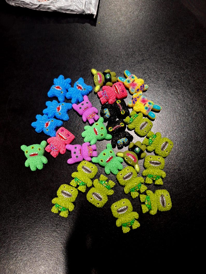 May include: A collection of colorful, miniature monster figurines in various colors, including blue, pink, green, yellow, and black. Each figurine has a unique design with open mouths and small details. The figurines are scattered on a dark surface.