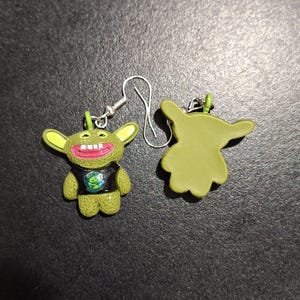 May include: A pair of novelty earrings featuring a green cartoon character with large ears, a wide grin, and a black shirt with a graphic. The earrings have silver-colored hooks.