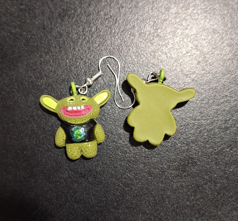 May include: A pair of novelty earrings featuring a green cartoon character with large ears, a wide grin, and a black shirt with a graphic. The earrings have silver-colored hooks.