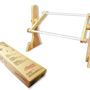 May include: A light-colored wooden embroidery frame stand with two horizontal white rods and adjustable side knobs. Beside it sits a rectangular cardboard box labeled American Dream Products and Tomorrow's Treasures with printed assembly diagrams.