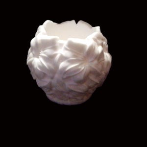 May include: A small, round, white vessel featuring a deeply embossed floral pattern covering the entire exterior surface. The object has a wide, open top with a scalloped rim and a smooth, matte finish, presenting a uniform white color throughout.