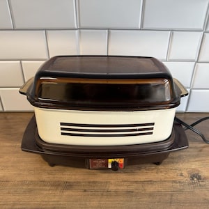 May include: A vintage brown and cream-colored electric slow cooker with a dark brown lid. The appliance sits on a brown base with a temperature control dial. The slow cooker has a rectangular shape with a cream-colored body and brown accents.