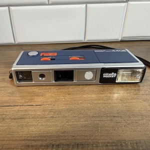 Puede incluir: Una cámara de película Minolta Pocket 440E azul y plateada con cuerpo rectangular, flash integrado en el lado derecho, visor e interruptores deslizantes naranjas en el panel superior. Una correa negra está sujeta al lateral del dispositivo.