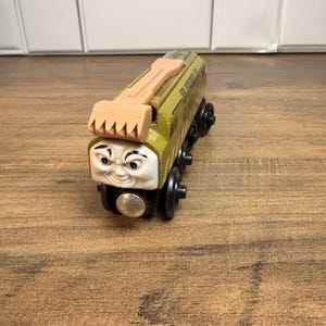 May include: A toy train engine featuring a tan excavator bucket attachment on top. The engine is olive green with a light-coloured face displaying a mischievous expression, black eyebrows, and black wheels with silver metallic centres on each side.