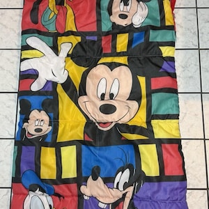 Puede incluir: Un saco de dormir rectangular con un patrón de cuadrícula colorido que incluye personajes de Disney como Mickey Mouse, Pluto, el Pato Donald y Goofy. El diseño utiliza bloques llamativos en rojo, amarillo, azul y morado con bordes negros.