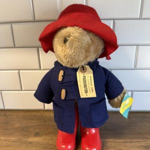 May include: A Paddington Bear plush toy wearing a red hat, a navy blue coat with wooden toggles, and red rain boots. The bear has a tag that reads "Paddington" and a small tag that reads "Eden".
