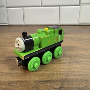 Könnte beinhalten: Eine grüne Spielzeugeisenbahn aus Holz mit einem lächelnden Gesicht an der Vorderseite, schwarzen Akzenten und der gelb aufgedruckten Nummer 11 an der Seite. Die Lokomotive hat sechs hellgrüne Räder und ein kleines gelbes Detail oben.