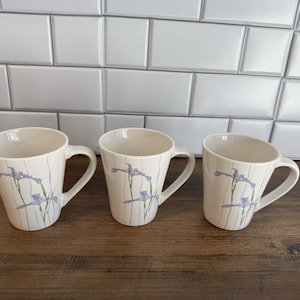 Puede incluir: Tres tazas de cerámica blanca con un diseño floral de iris moradas y tallos verdes. Las tazas tienen forma curva y asa. El fondo es una pared de azulejos blancos y una superficie de madera.