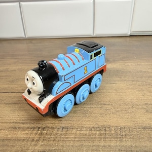 Puede incluir: Una locomotora de juguete azul con frente negro y cara blanca, detalles rojos en la base y el número 1 impreso en amarillo en el lateral. El tren cuenta con seis ruedas azules y un techo negro, diseñado como una clásica locomotora de vapor.