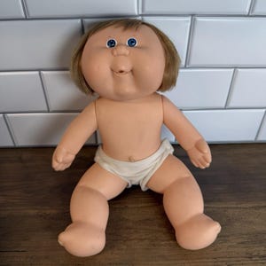 May include: A vintage baby doll with a round face, blue eyes, and short brown hair. The doll is wearing a white diaper and has a smiling expression. The doll is made of a flesh-toned material.