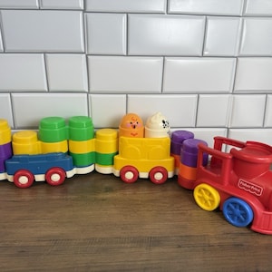 Puede incluir: Un colorido tren de juguete de plástico que incluye una locomotora roja con el logotipo de Fisher-Price, conectada a varios vagones. Los vagones están construidos con bloques apilables amarillos, verdes, morados y azules con dos figuras.